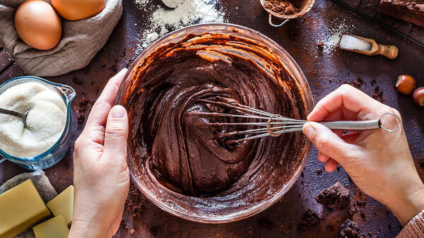 Hands mix chocolate batter with a mixer, cake ingredients are laid out around them.