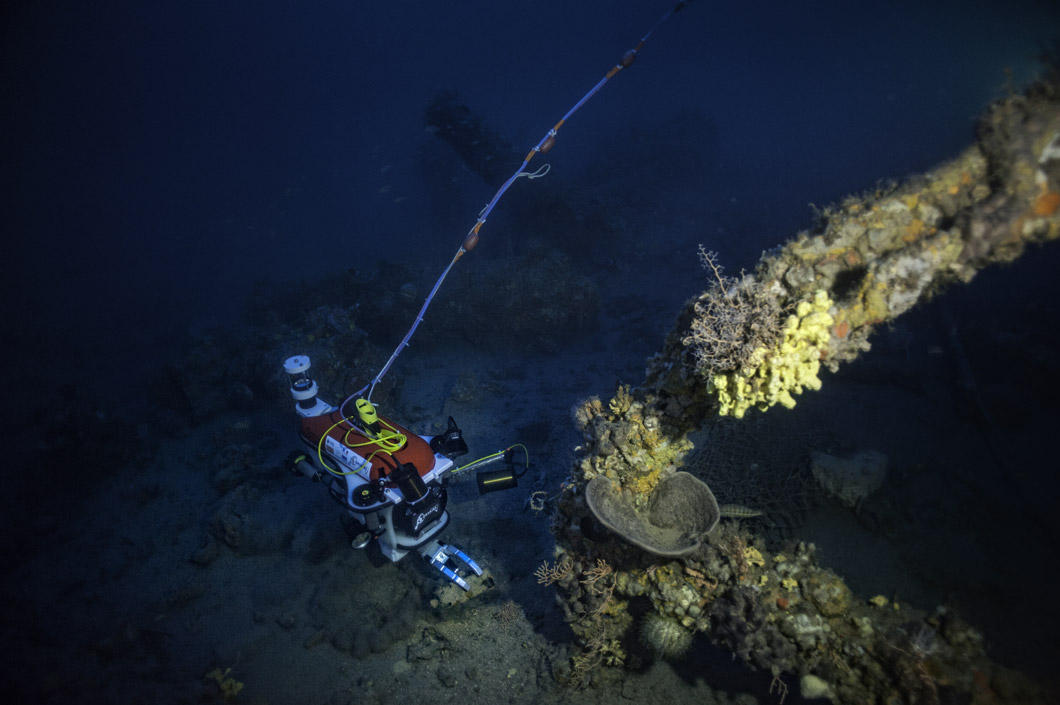 The robot is on the sea floor. In the foreground: a boat anchor. A cable is visible attached to the robot.