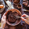 Hands mix chocolate batter with a mixer, cake ingredients are laid out around them.